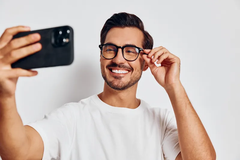 Smiling man taking a selfie wearing glasses, casual white t-shirt, looking cheerful and confident at camera against plain solid light background. Lifestyle concept.