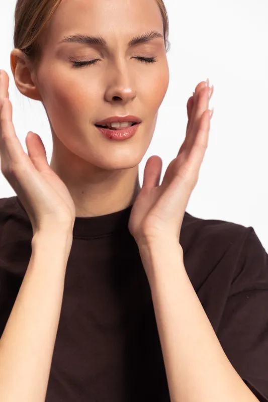 Young woman with closed eyes touching her face with both hands, wearing a dark top, isolated on white background. Concept of skincare, relaxation, and natural beauty care.