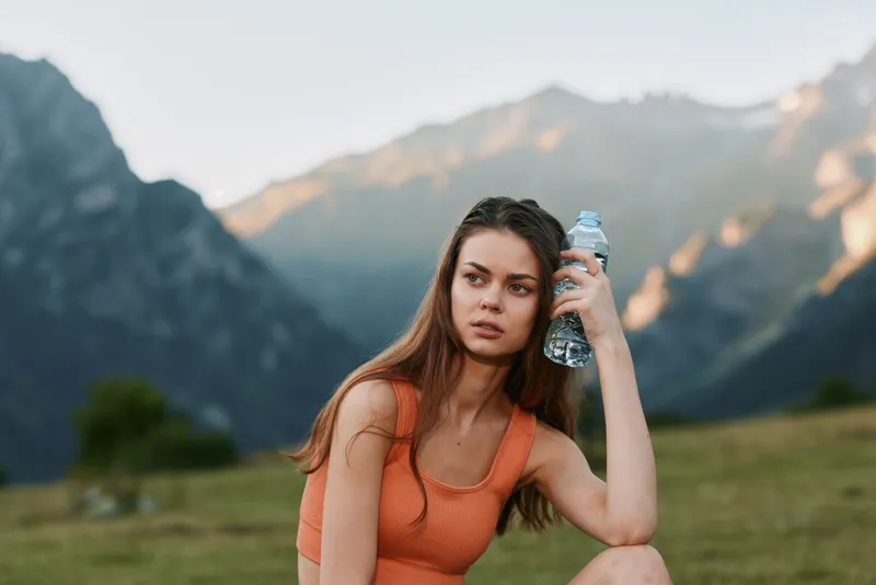 A young woman sits outdoors in a mountainous landscape, wearing an orange sports top and holding a water bottle. She gazes into the distance with a calm, focused expression.