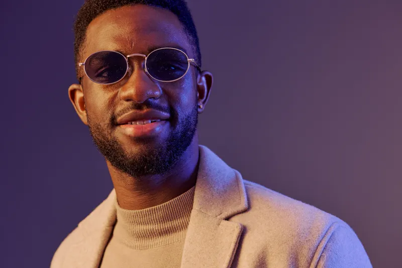 Stylish young man with sunglasses smiling, wearing a beige sweater and jacket, set against a colorful background, exuding confidence and modern fashion