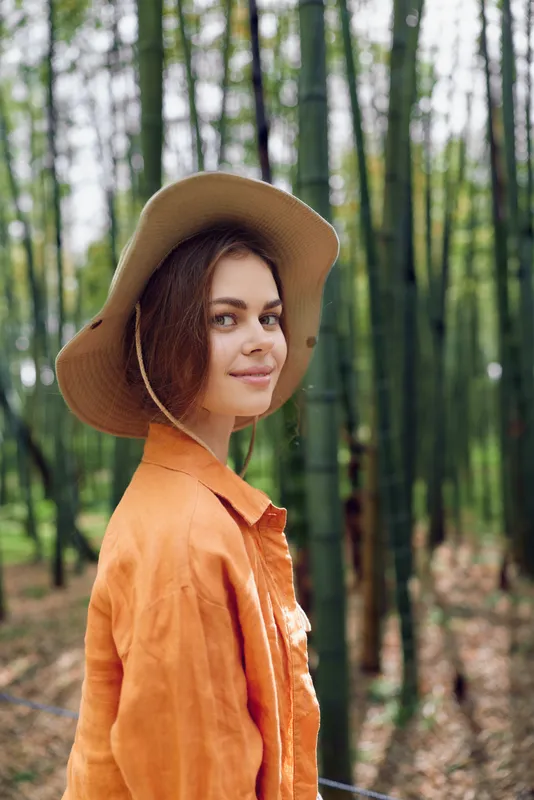 Woman in a straw hat smiles in a bamboo forest portrait outdoors, nature background. Female model in orange shirt and jacket, serene lifestyle scene with green trees and stalks.
