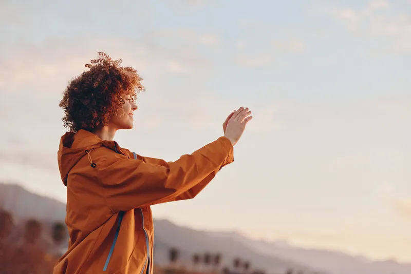 Woman lifestyle outdoors at sunset, curly hair in profile, wearing brown jacket, raising one hand to wave, standing on hill with distant mountains, soft sky, calm mood