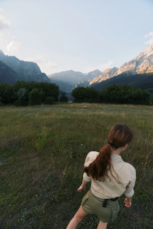 Young woman with long brown hair walks through a grassy field surrounded by mountains during a clear day, enjoying a peaceful outdoor lifestyle moment.