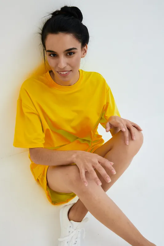Young woman wearing bright yellow dress and white sneakers posing on white background, smiling with natural makeup and casual hairstyle, modern minimalistic style.