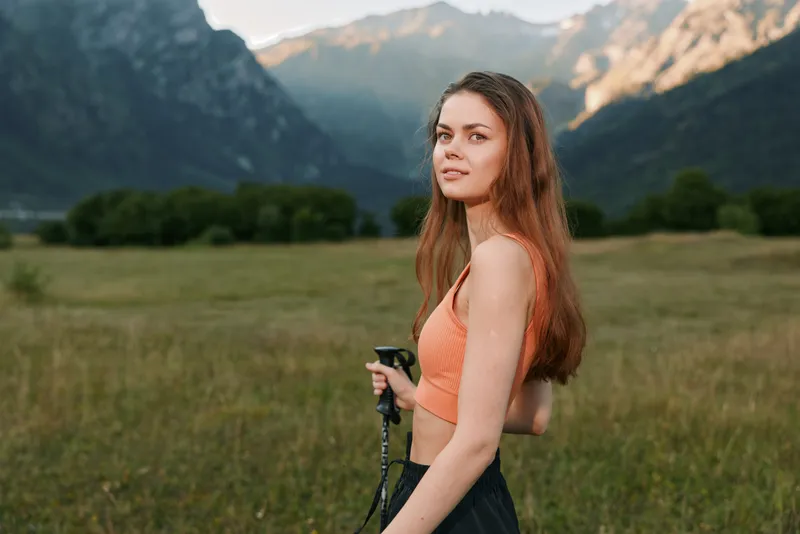 A young woman stands in a grassy meadow with distant mountains, wearing an orange crop top and dark athletic pants, holding trekking poles, looking toward the camera.