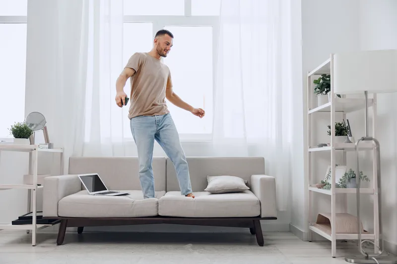 Energetic man dancing on a couch, showcasing a joyful moment in a bright living room filled with natural light and modern decor. Perfect for lifestyle themes.