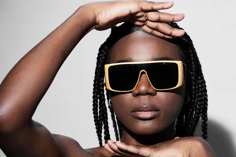 Stylish young Black woman wearing oversized sunglasses poses confidently with hands framing her face against a minimalistic background, showcasing contemporary fashion