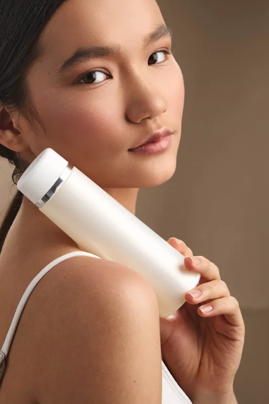 Asian woman holds a sleek skincare bottle in a soft studio setting, close portrait with calm expression, emphasizing personal care and modern wellness.