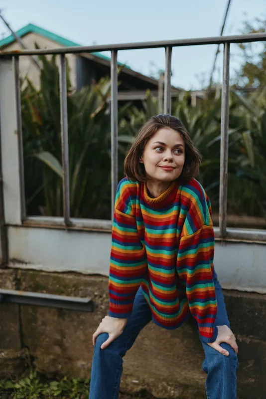 Woman portrait sweater colorful outdoors casual sitting smile leaning on fence in backyard, candid fashion portrait with jeans and short hair, natural light and relaxed cozy vibe.