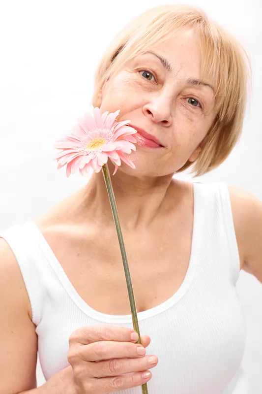 Elderly woman portrait, holding pink gerbera flower near face, blonde hair, white studio background, soft smile, bright light, calm minimal composition, gentle focus