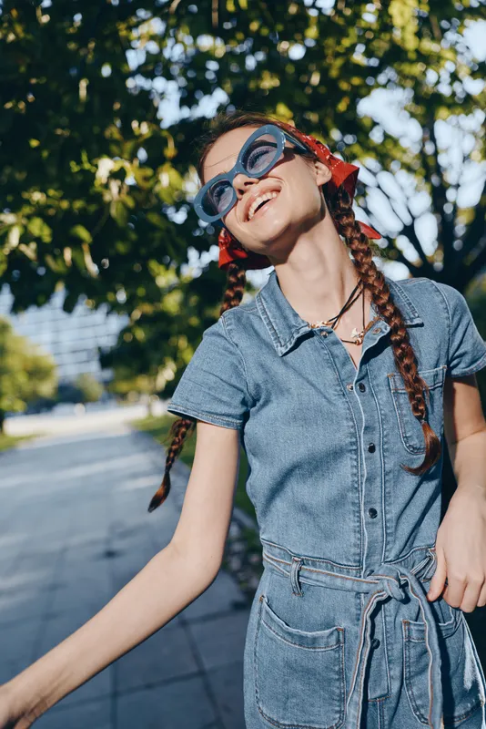 Lifestyle woman in modern boho-western denim street style enjoying outdoor sunshine with braided hair and retro sunglasses, film color aesthetic.