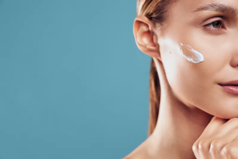 Close up of young woman with clear skin applying moisturizing cream on cheek against blue background, skincare routine concept focused on beauty and healthy skin care.