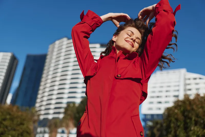 Joyful woman outdoors in a red coat, smiling under bright sun, wind in her hair, urban backdrop, clear blue sky, sharp lighting, relaxed pose, lush city energy.
