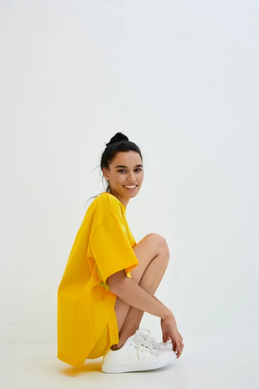 smiling woman wearing bright yellow oversized t-shirt and white sneakers, casual fashion, minimalist white background, youthful and cheerful expression