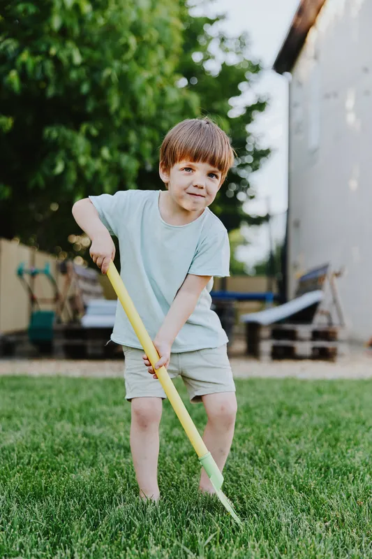 Playful boy enjoying outdoor activities, wearing a light blue shirt and shorts, smiling while using a yellow shovel in the garden, surrounded by green grass.