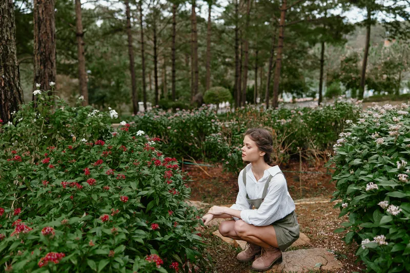 Young woman in green shorts and white blouse squatting among colorful flowers in a serene forest garden, depicting harmony with nature and tranquility