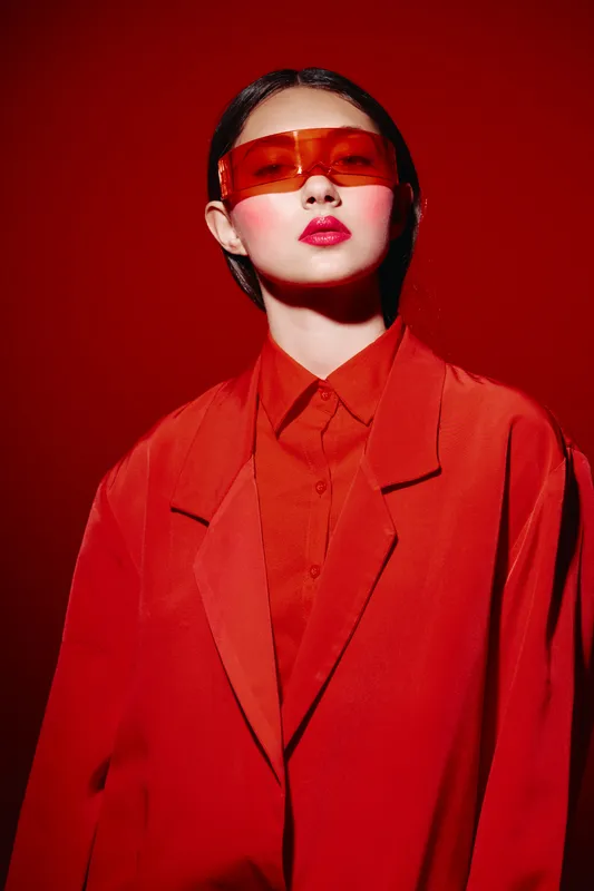 Fashion model in oversized orange blazer and matching shirt poses against red background. sunglasses and bright lipstick emphasize creative bold beauty concept in studio lighting.