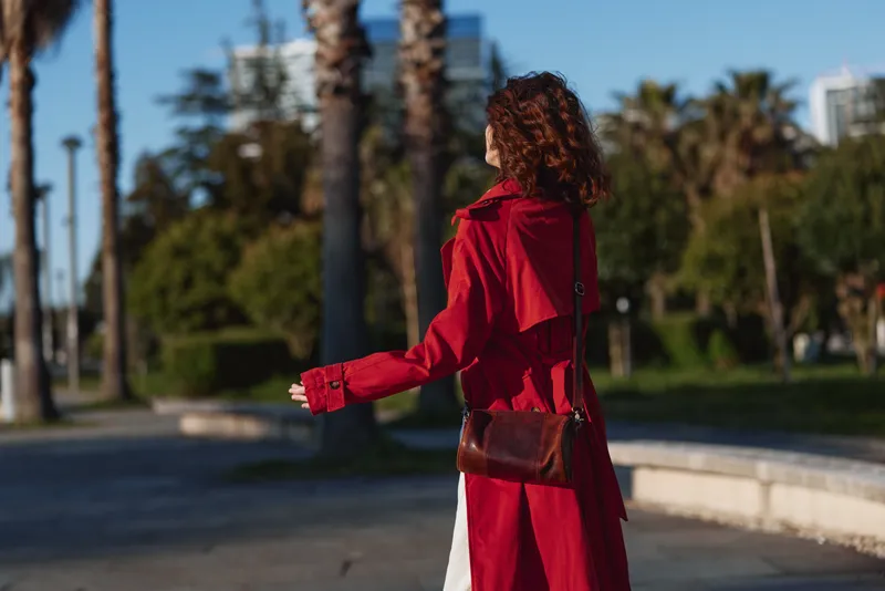 A woman in a red coat walks outdoors in a sunny urban park, carrying a brown bag. Fashion mood, movement, and casual street style. Her silhouette stands out against palm trees and modern buildings,