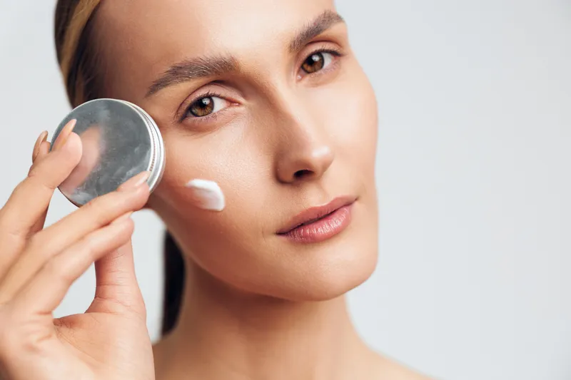 Skincare woman face cream closeup wellness beauty treatment, applying white cream from a silver jar on smooth skin. Soft studio light on neutral background, self care mood calm ritual.