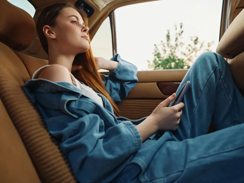 Young woman dressed in denim relaxes in a car, enjoying her phone while gazing out of the window, conveying a sense of serenity.