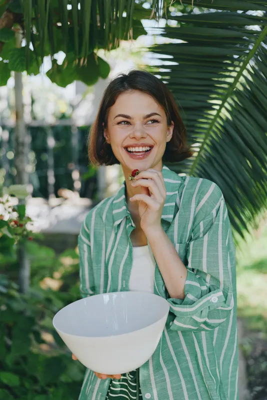 Smiling woman outdoors holds a white bowl, enjoying a sunny garden moment in a green striped shirt and casual attire.