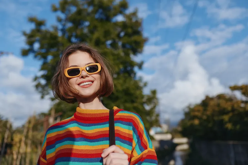 Young woman in a striped sweater and yellow sunglasses smiling in an outdoor park. Short bob haircut, holding bag strap, casual outfit and sunny travel lifestyle portrait.