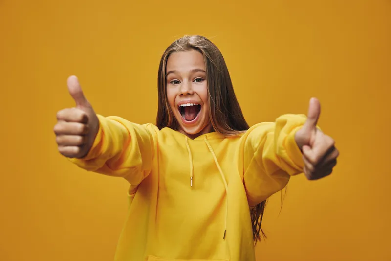 Girl in yellow hoodie shows thumbs up with smile and excitement on orange background. Young person with long hair expressing positivity and happiness indoors with casual style.
