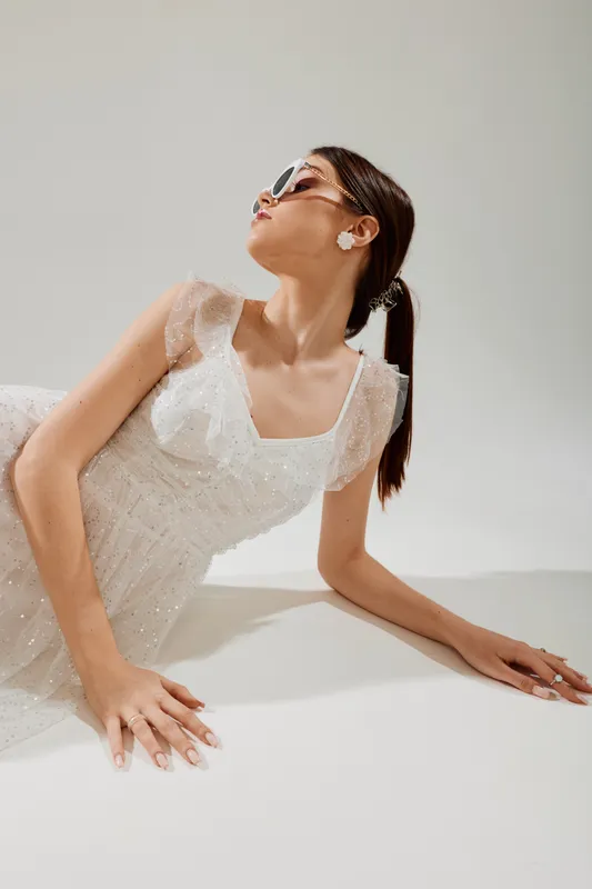 Young woman in a white lace dress poses with sunglasses and earrings against a plain background. Concept of woman beauty fashion and elegant style.