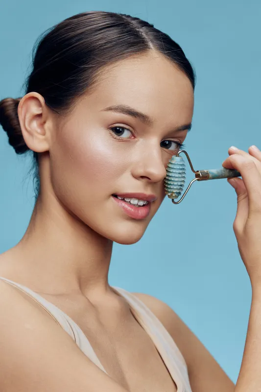 Young woman using a jade roller for skin care on a blue background, emphasizing beauty, relaxation, and healthy lifestyle