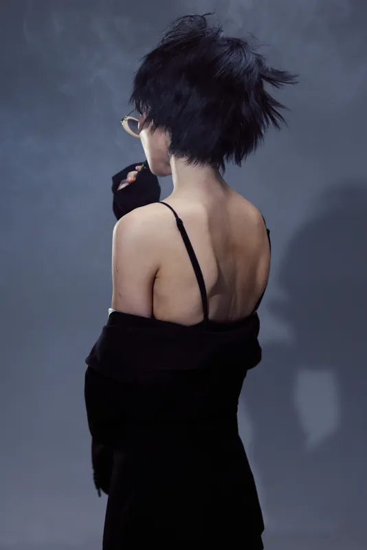 Woman with short black hair and glasses wearing black clothing turned back on smoky background