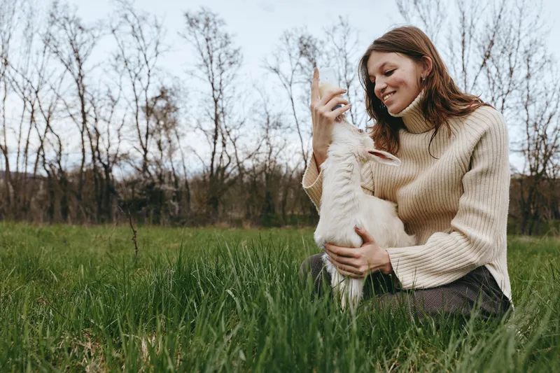 Smiling woman in a knit sweater sits in a grassy field, bonding with a playful goat kid. Soft natural light and relaxed posture create a warm lifestyle moment focused on friendship and outdoor nature.