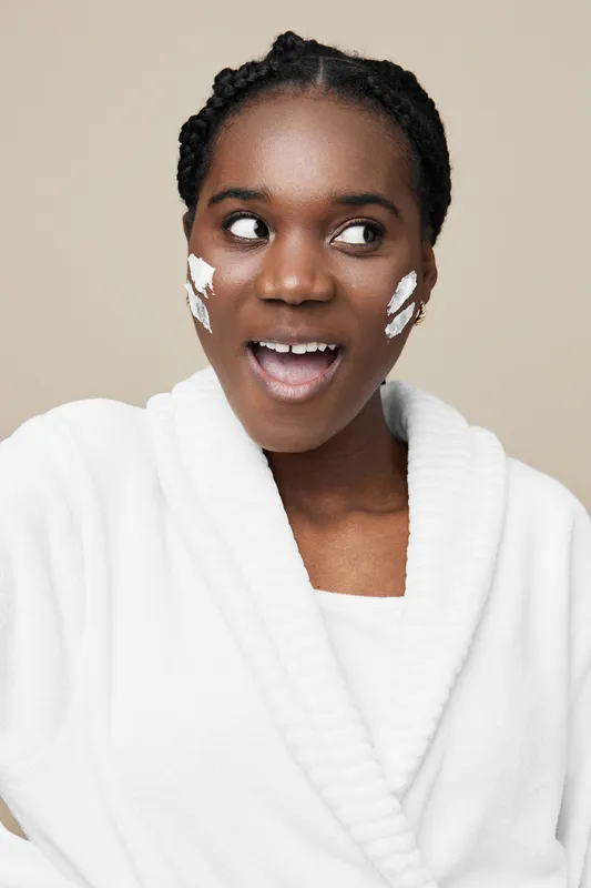 Black female model with a cheerful expression applying skincare products, wearing a white bathrobe against a neutral beige background