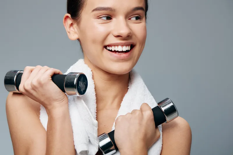Fitness woman lifting dumbbells with a towel draped over her shoulders, smiling during workout in a studio, showcasing strength, balance and personal wellness.