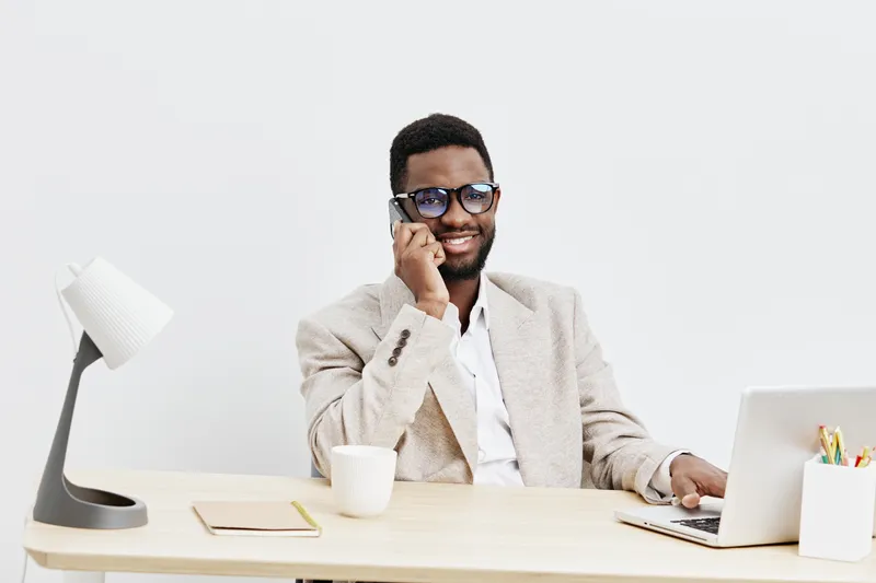Confident young smiling African American man in beige blazer and sunglasses working at modern desk with laptop isolated on plain white background studio portrait. Business lifestyle concept