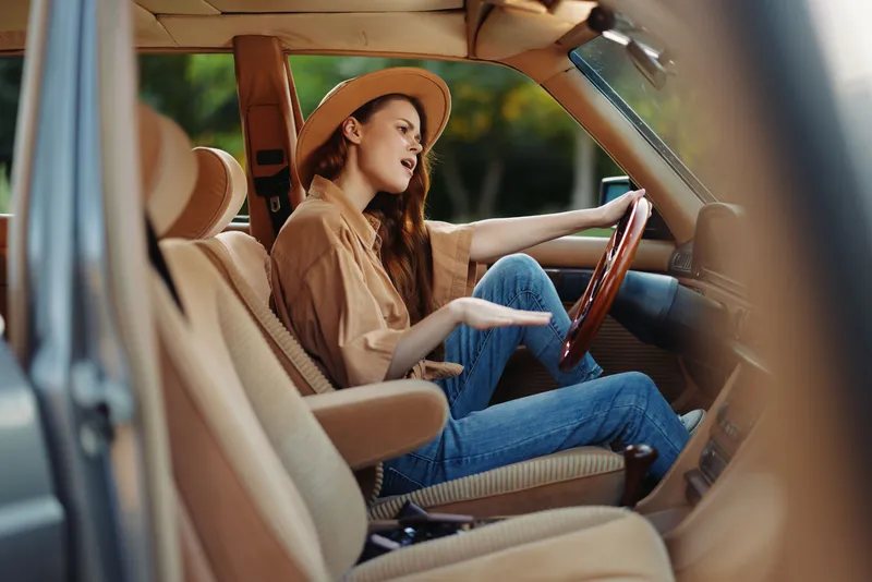 Young woman in casual outfit enjoying a scenic drive, expressing emotions while sitting in a classic car with a stylish hat.