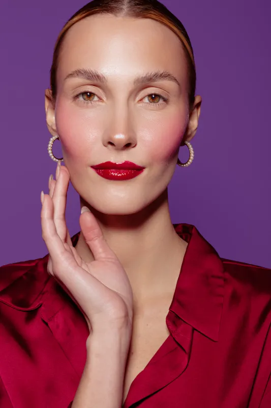 Portrait of a confident woman with red lipstick and hoop earrings wearing a red satin blouse on purple background. Concept of beauty, fashion, elegance, and makeup.