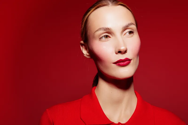 Portrait of a woman with red lipstick and red shirt on red background. Soft lighting highlights her clear skin and elegant expression. Concept of beauty, fashion, and style.