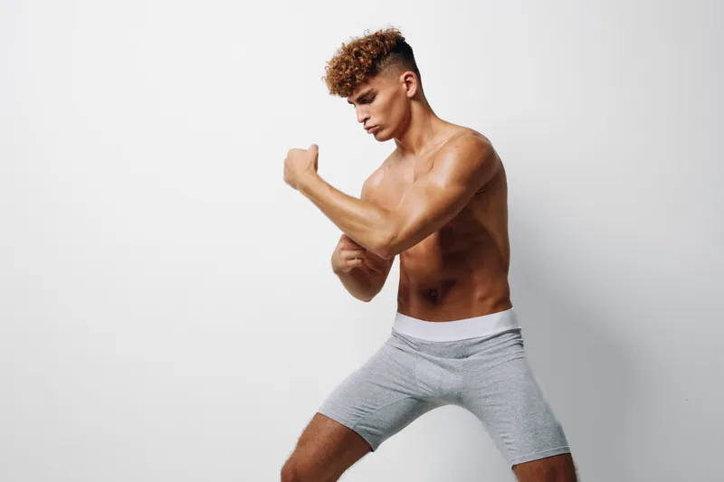 Athletic young man, shirtless in gray shorts, standing in a boxing stance for fitness training and workout against white wall. Muscular body and concentration for exercise or sport.