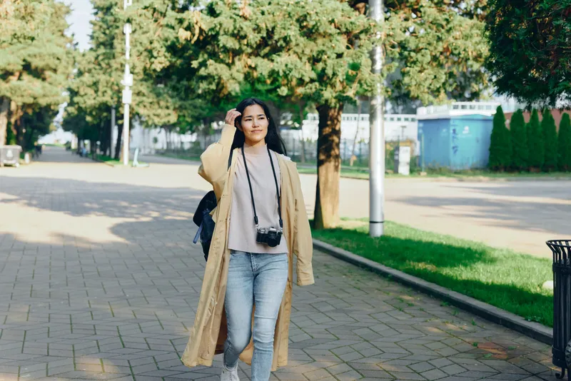 Woman walking along a park path in a long beige coat, camera hanging from neck, casual street fashion, daytime light, urban greenery, candid lifestyle photo for travel or style blogs