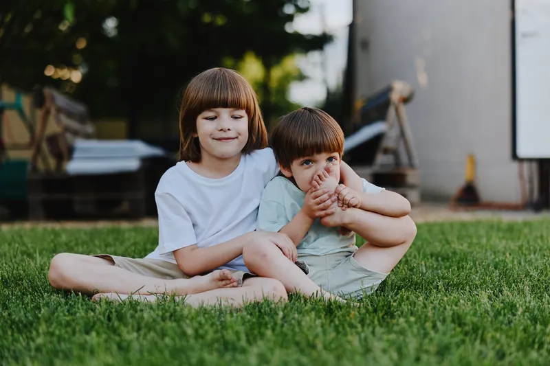 Two happy children enjoying a sunny day on the grass, one boy playfully teasing the other with a smile, creating a joyful atmosphere of childhood exploration.
