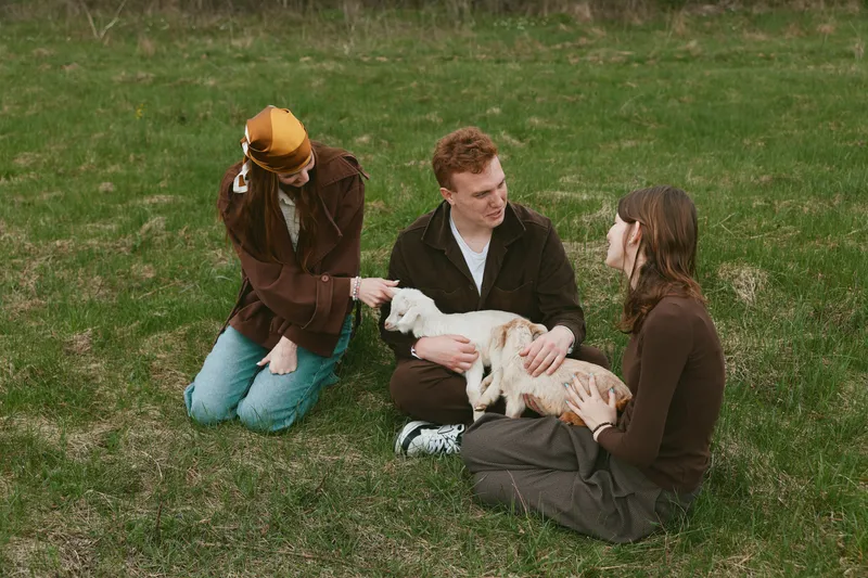 Goat, kid, friends, outdoor, park, petting, animal, family describe a candid lifestyle scene as three people softly interact with a gentle goat kid on a grassy field, smiles, relaxed clothing,
