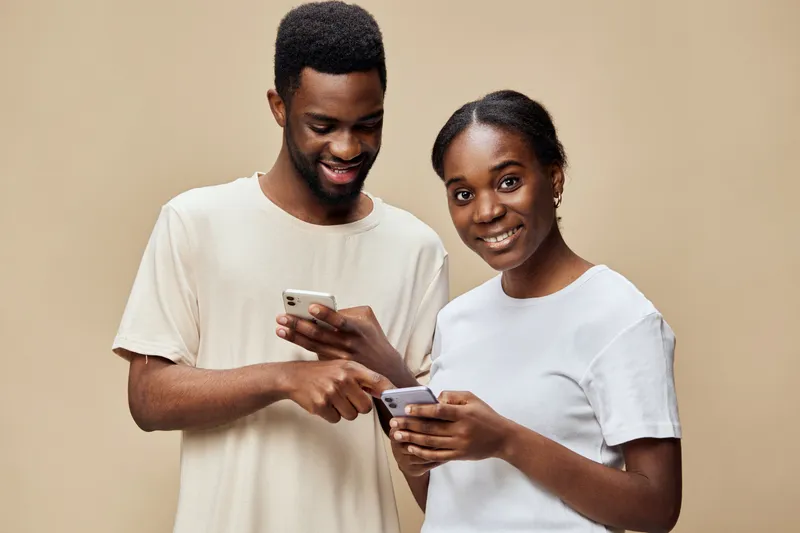 Young couple engaged with smartphones, sharing content and smiling, showcasing modern technology and connection in a neutral toned background