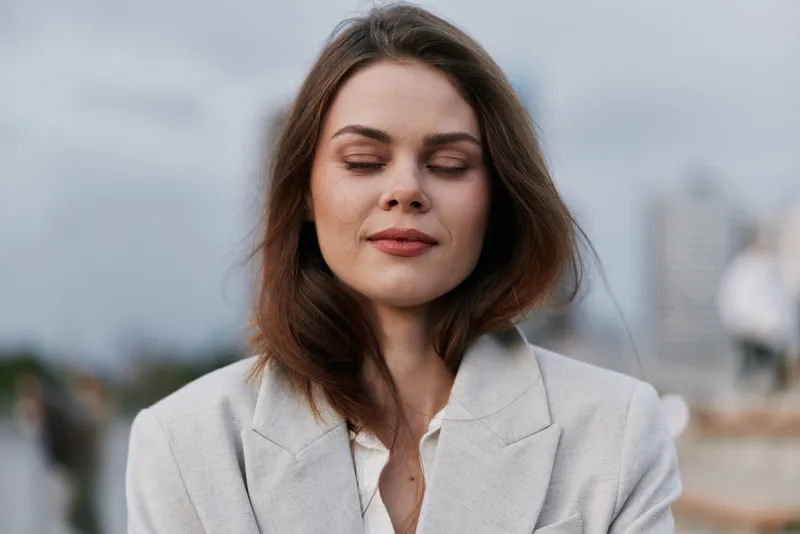 Calm woman with closed eyes in beige blazer outdoors showing peaceful expression and natural makeup in soft daylight with blurred urban background