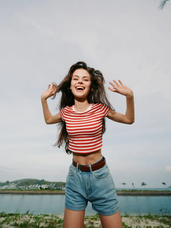 Joyful young woman outdoors in a striped red and white tshirt and denim shorts, arms raised, bright smile, capturing carefree energy and sunny summer mood.