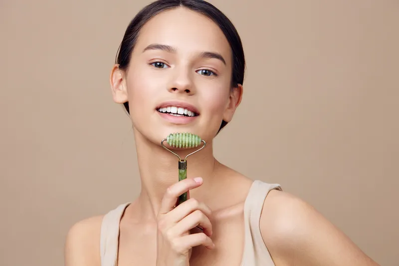 Young woman with smooth skin using a jade roller for skincare, showcasing a natural beauty routine in a minimalistic beige background