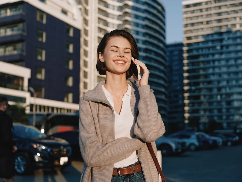 A cheerful urban professional speaks on a mobile phone in a city setting. She smiles warmly, dressed in a light coat and casual attire, projecting a confident, friendly vibe.