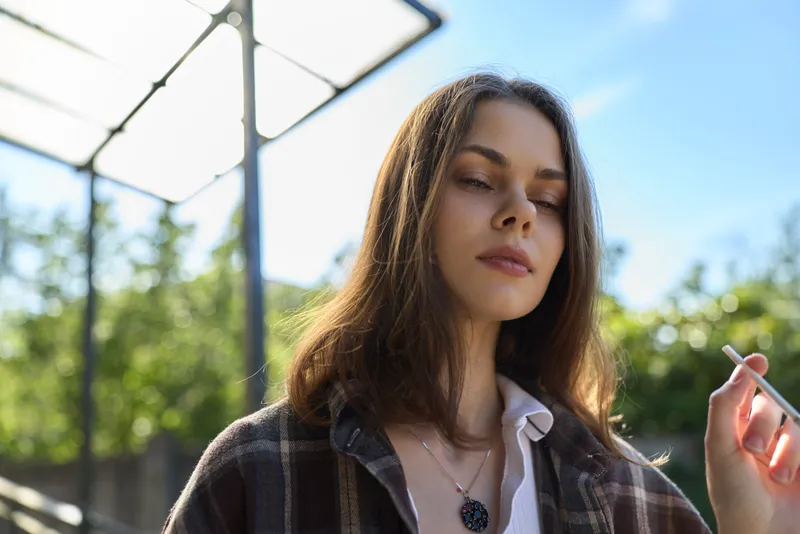 Casual woman lifestyle portrait outdoors with sunlit background, relaxed expression, plaid shirt, open collar, delicate necklace, calm mood, candid moment