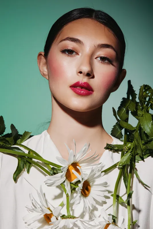 Portrait of a young woman with bold red lips and natural makeup, wearing a white shirt with fresh daisies. Fashion creative concept with a clean green background.