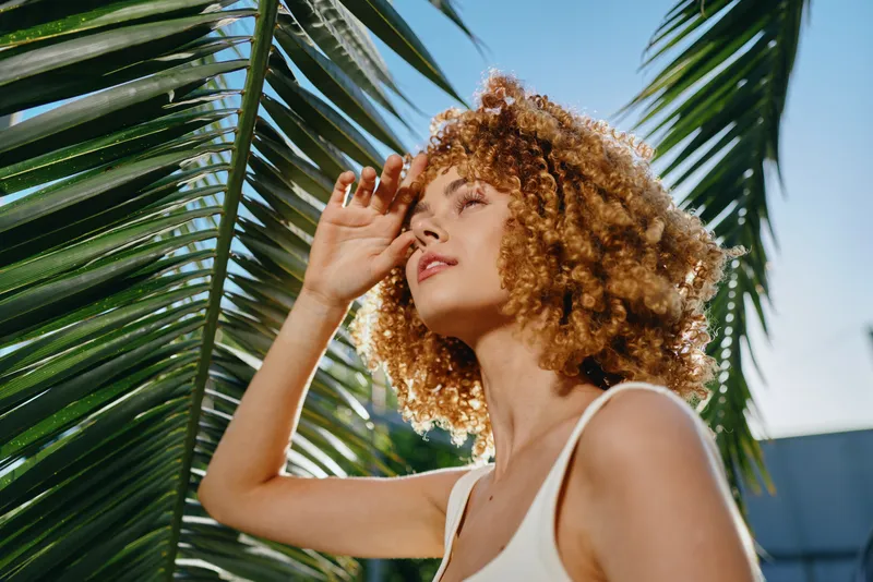 curly woman enjoying sunlight under palm leaves, with natural makeup and casual white top, capturing a bright and sunny summer mood outdoors.