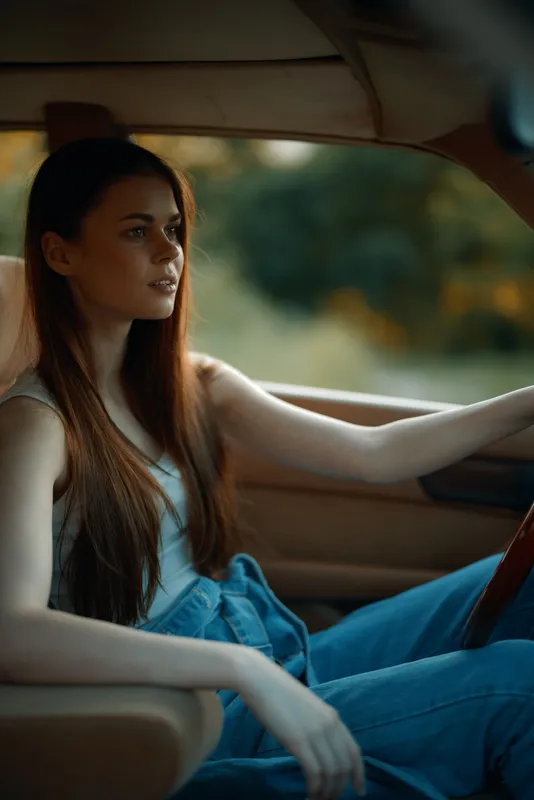 Young woman with long hair sitting in a vintage car, wearing a casual outfit, looking thoughtfully out of the window. The warm light creates an autumn atmosphere.